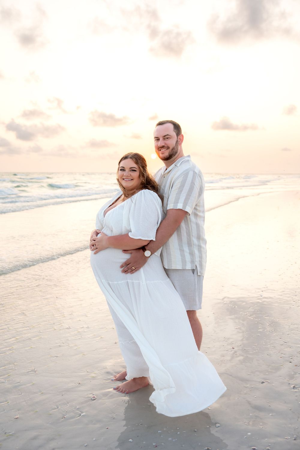 Smiling pregnant couple embracing on a sunlit beach with ocean waves behind them.