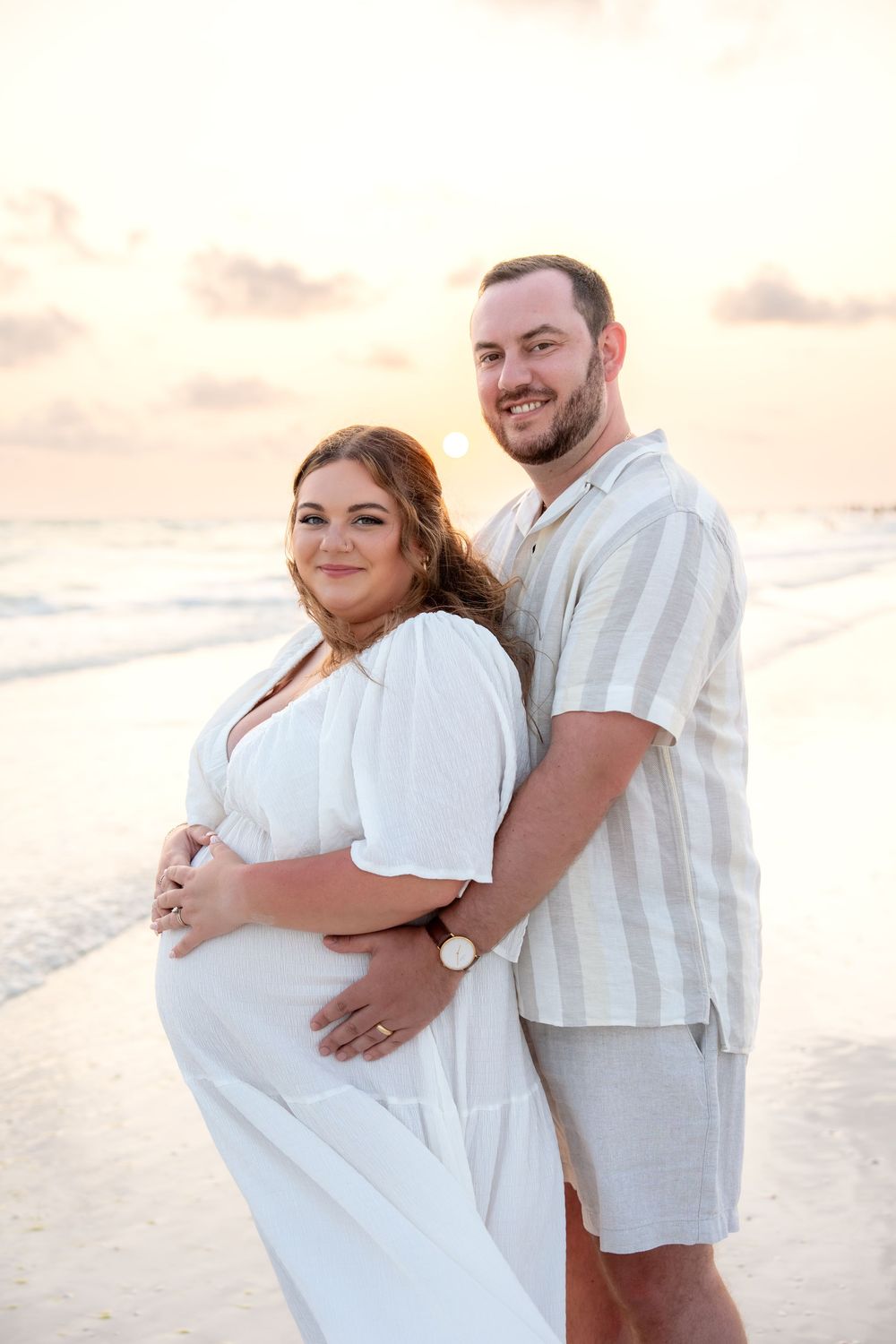 Happy expecting couple posing together on beach at golden sunset with ocean backdrop.