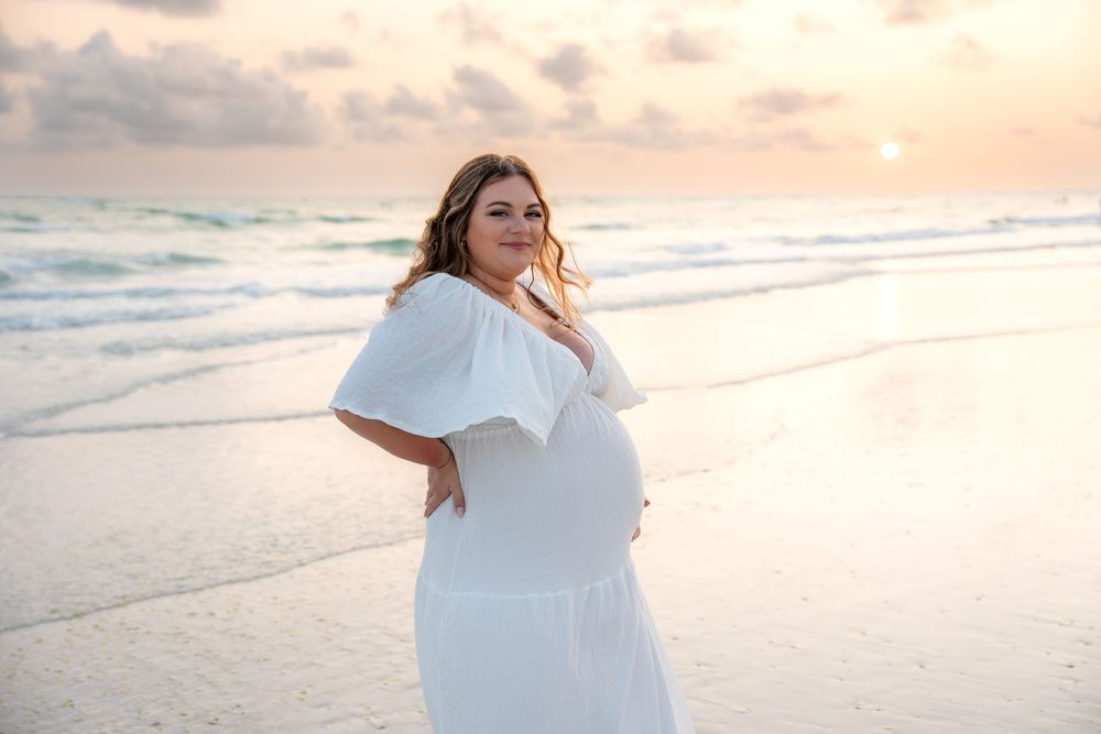 Pregnant woman in white off-shoulder dress posing gracefully on beach at sunset.