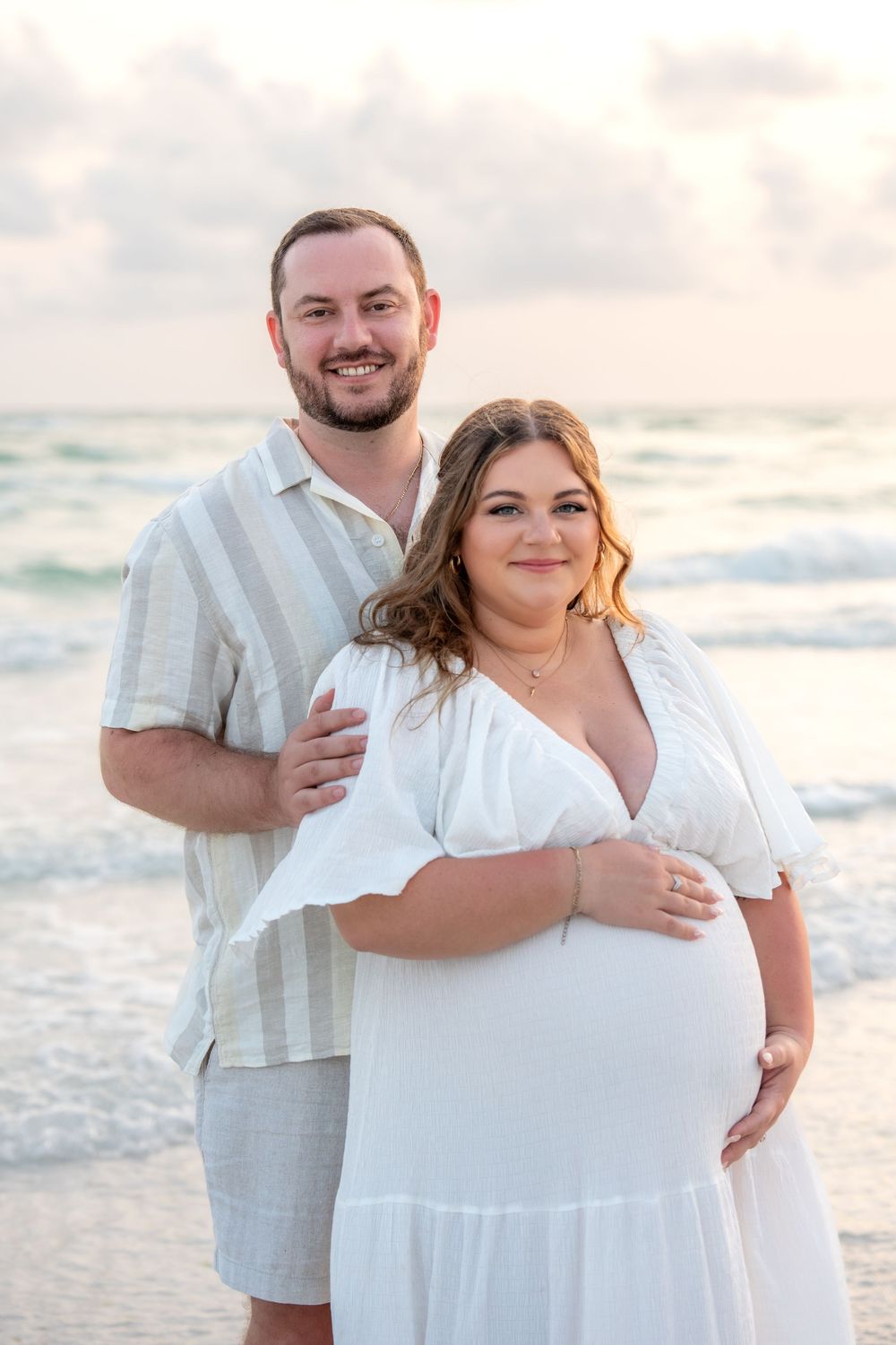 Smiling maternity couple standing together on beach with turquoise ocean at sunset.
