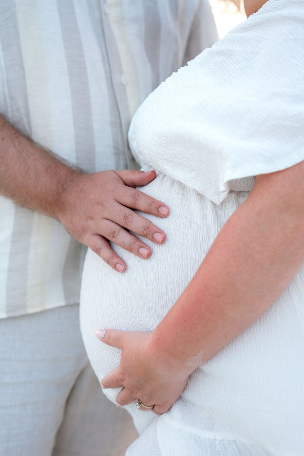 Close-up of couple's hands gently cradling pregnant belly on beach, woman wearing engagement ring.