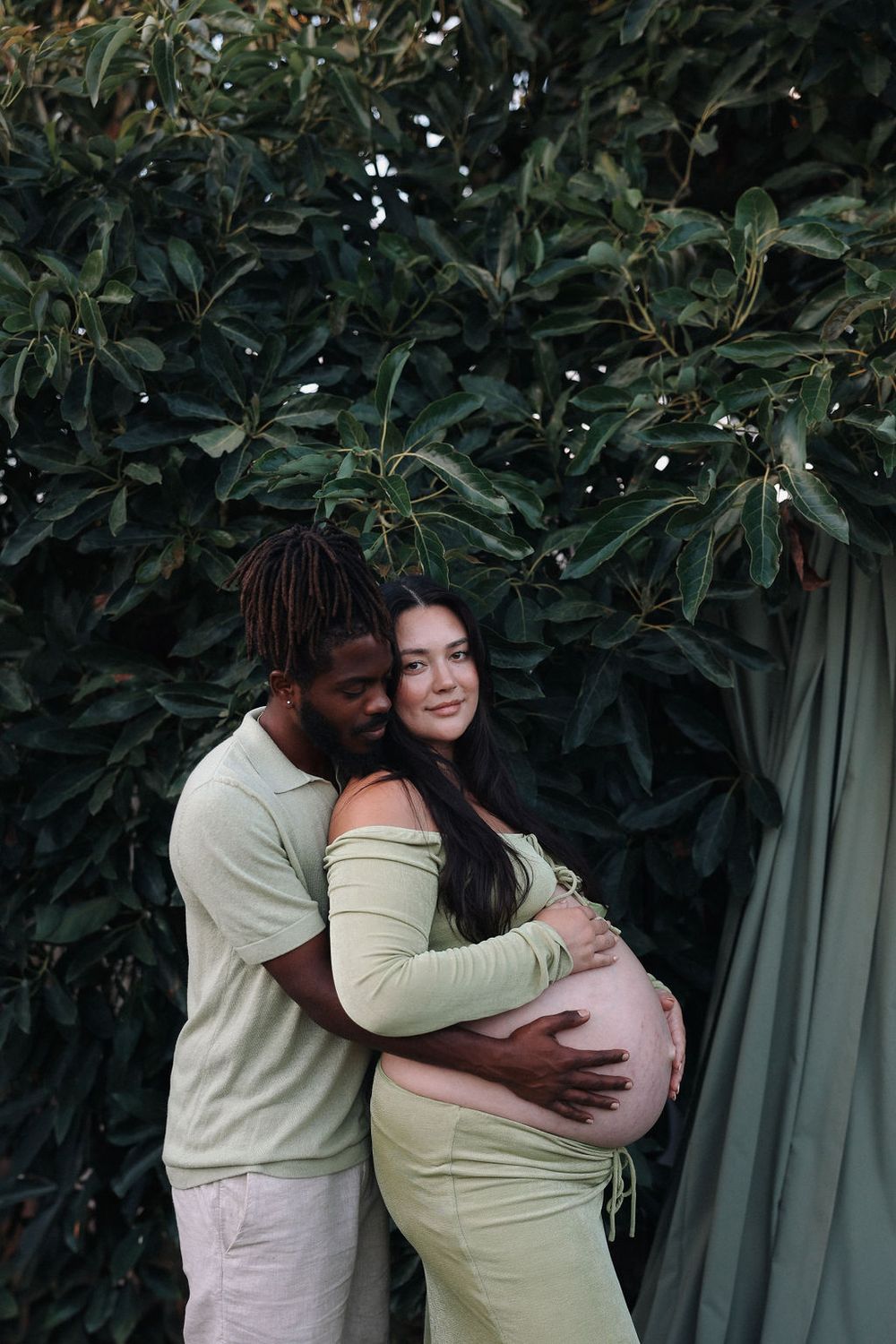 A couple shares a tender embrace against a backdrop of lush green foliage while wearing light colored clothing.
