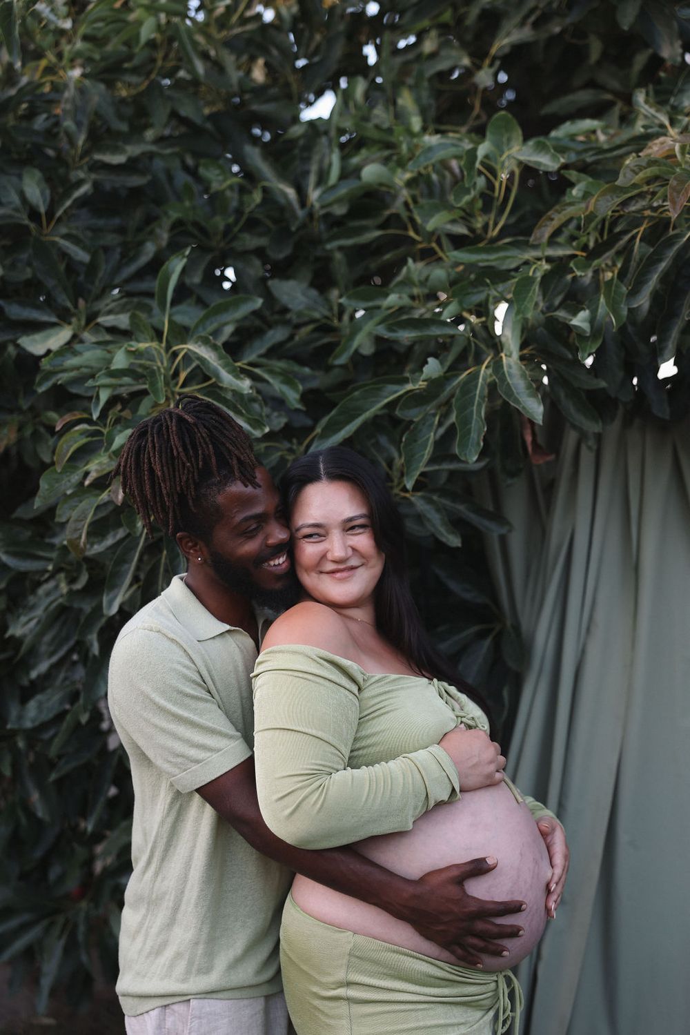 An expecting couple embraces lovingly in front of green foliage, sharing a tender moment in matching light-colored outfits.