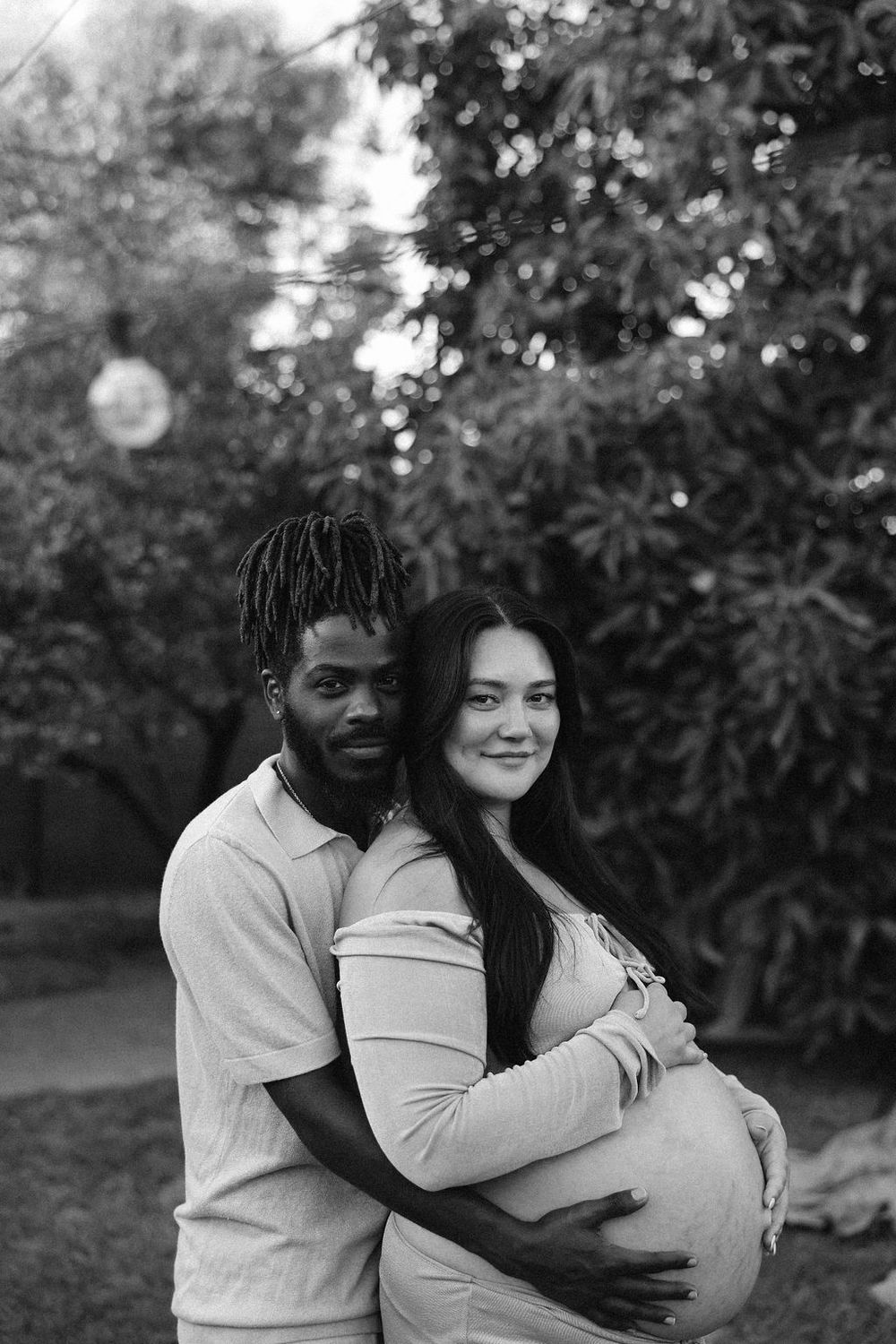 A black and white portrait of an expectant couple sharing an intimate embrace outdoors with trees in the background.
