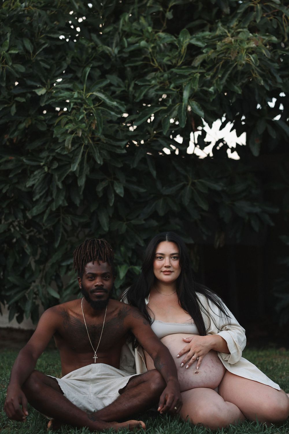 A couple poses together outdoors against green foliage in matching light colored outfits during a photoshoot.