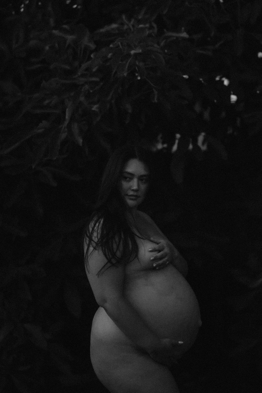 Black and white artistic portrait photographed in a dark outdoor setting with trees and shadows.