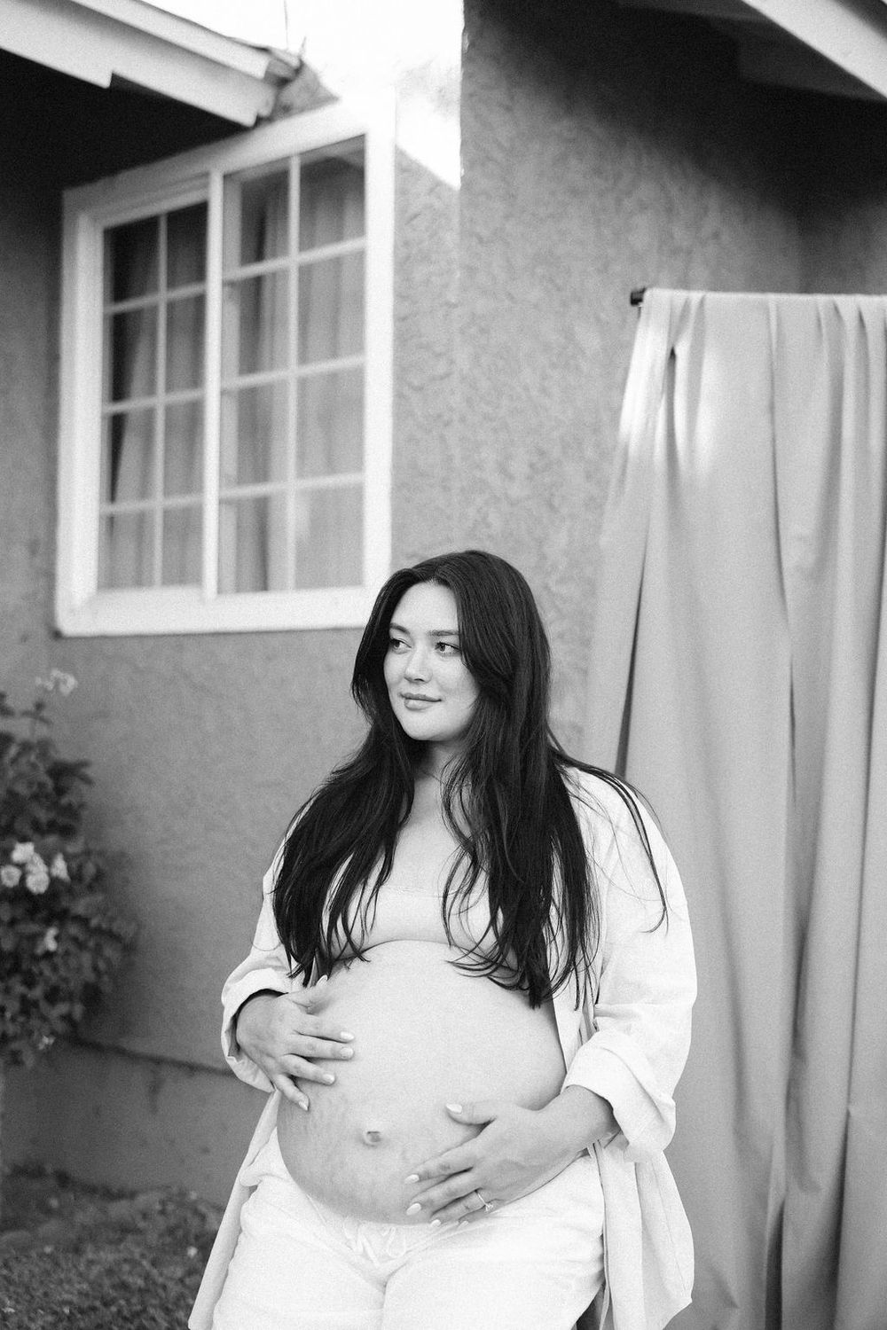 A black and white portrait of an expectant mother in a white shirt standing near a house window.