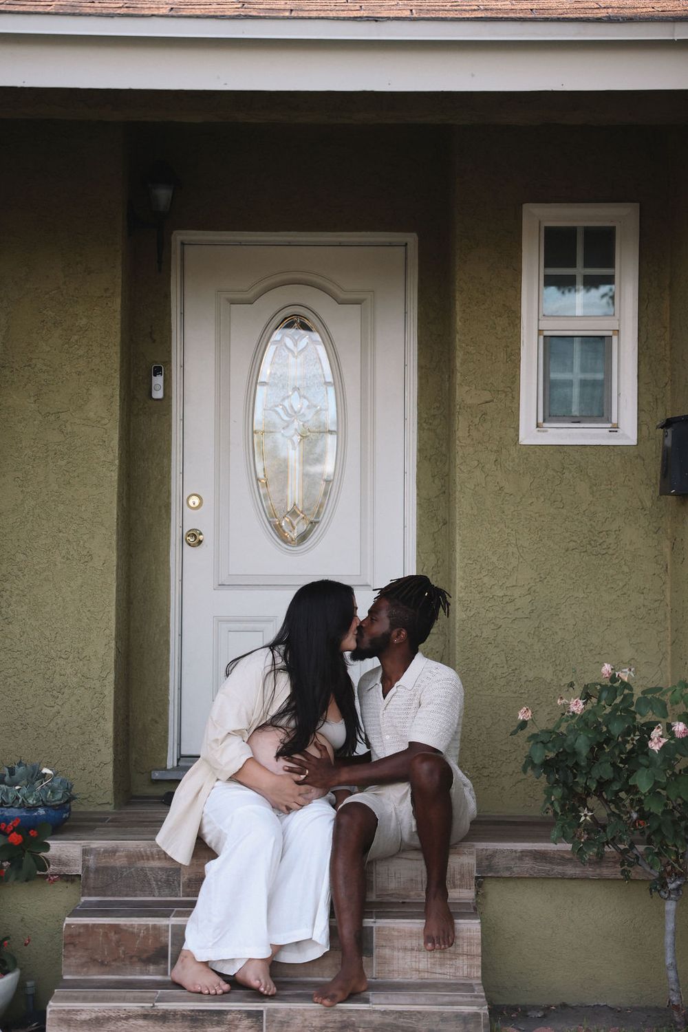 A couple sits together on the front steps of a house with olive stucco walls and a white door with an oval window.