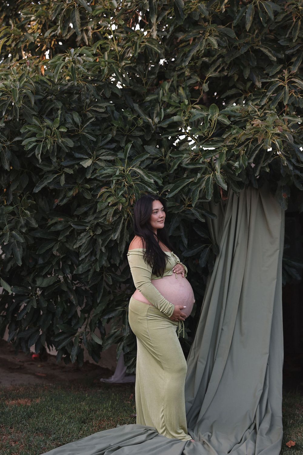 A person in a yellow dress poses against a backdrop of green foliage and gray curtains in a garden setting.