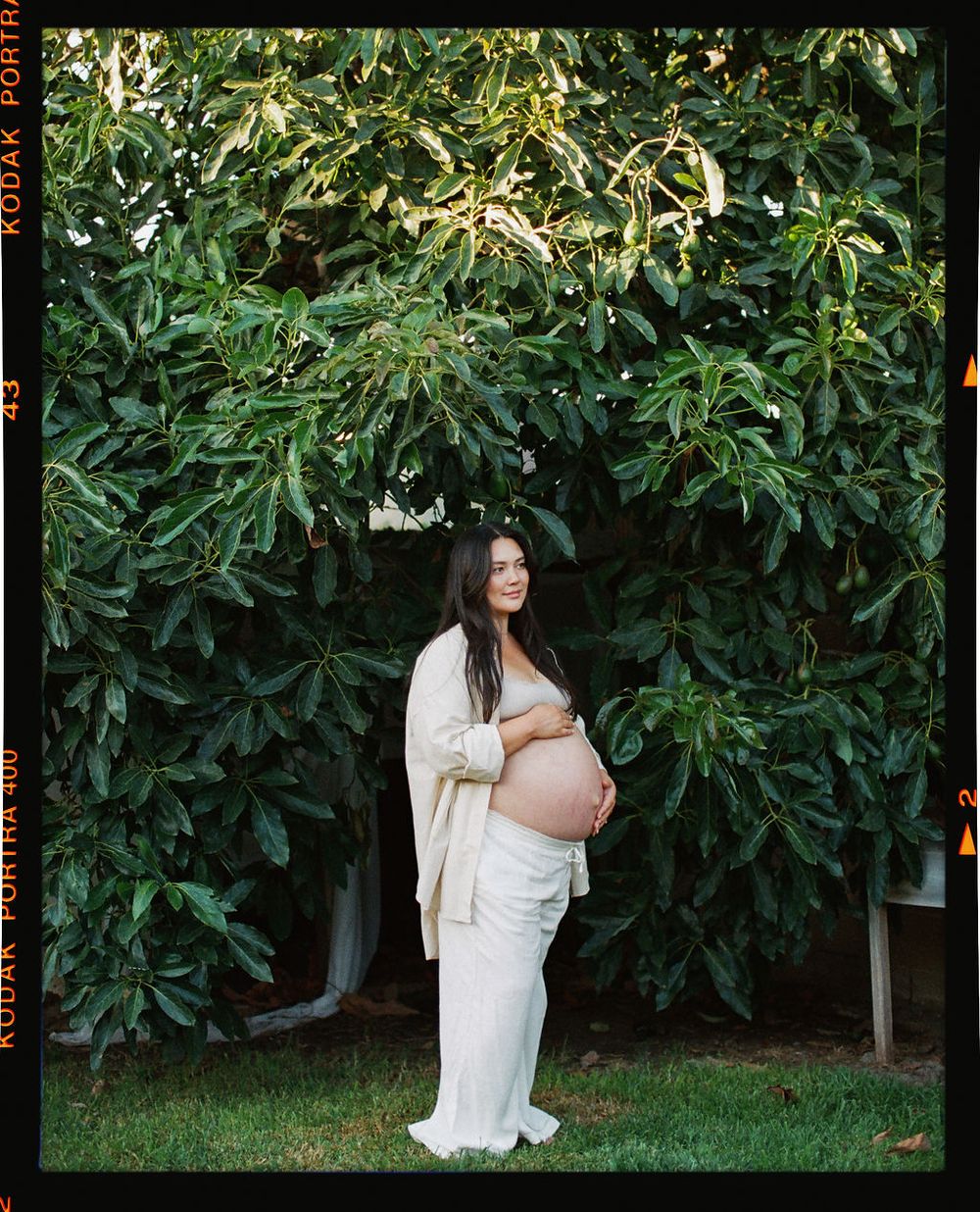 A pregnant woman in a flowing white dress stands in a garden with lush greenery and foliage behind her.