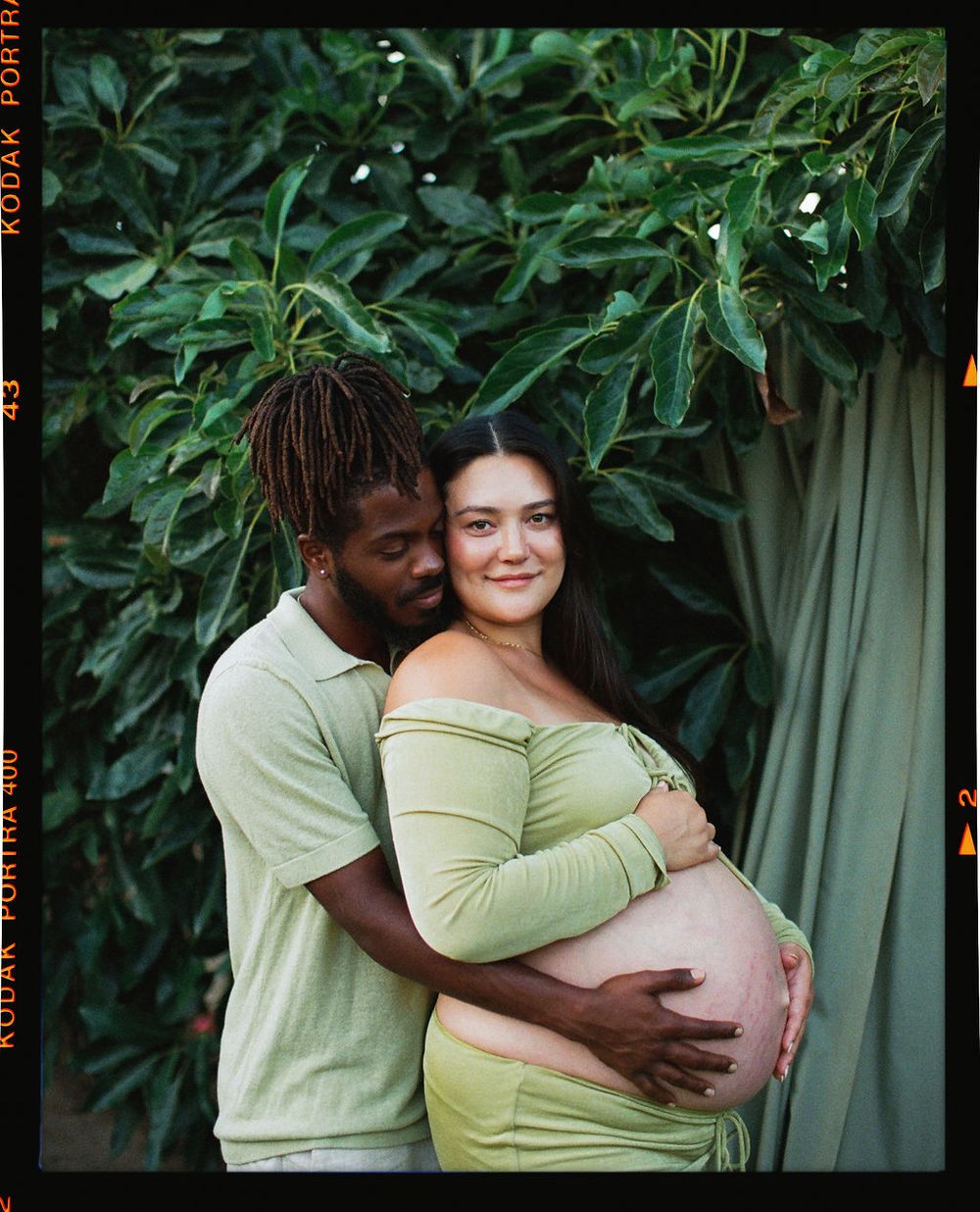 A couple embraces near lush green foliage while celebrating their upcoming arrival with a maternity photo.