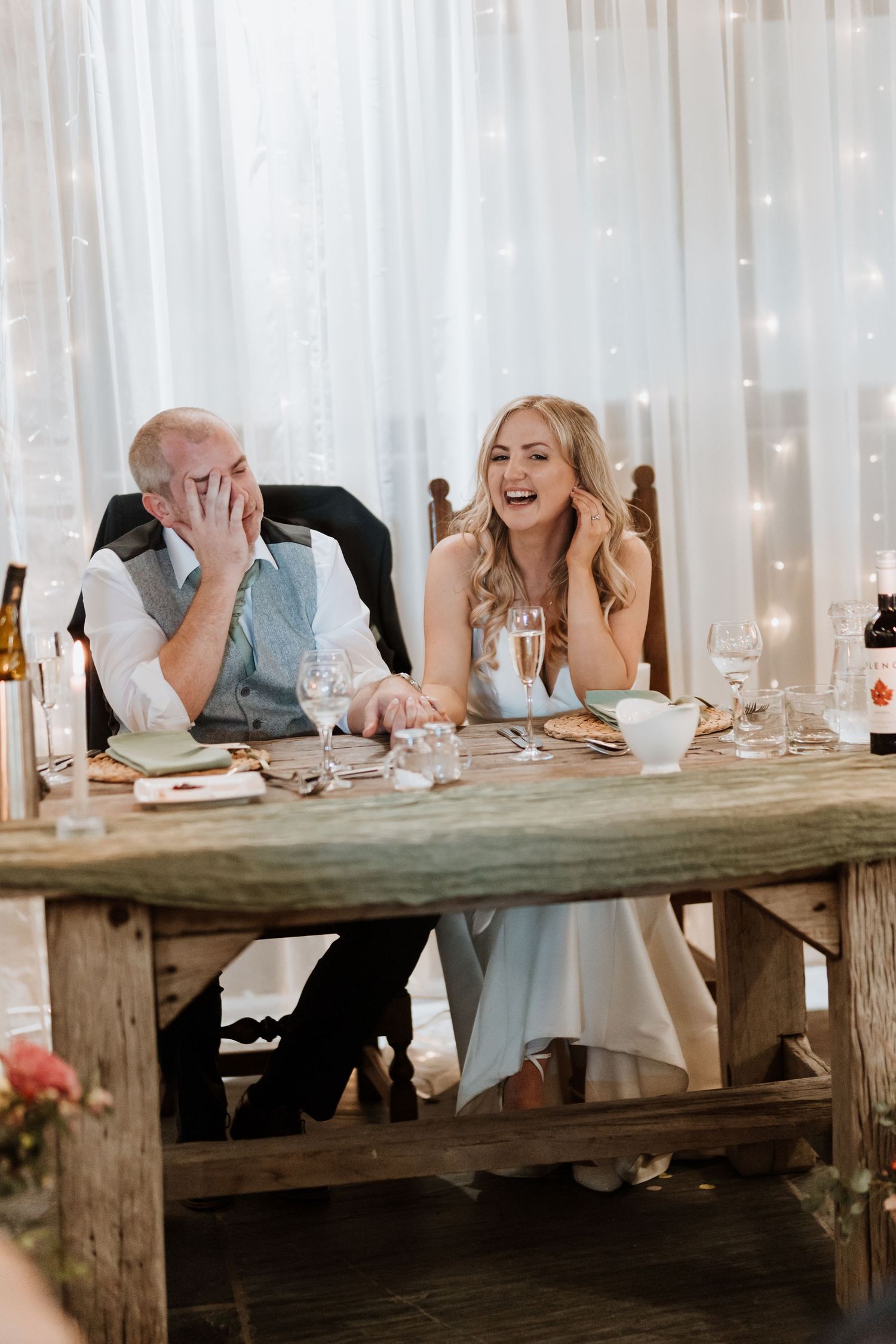 A couple shares a candid laugh while seated at a rustic wooden table during their wedding reception with twinkle lights behind them.