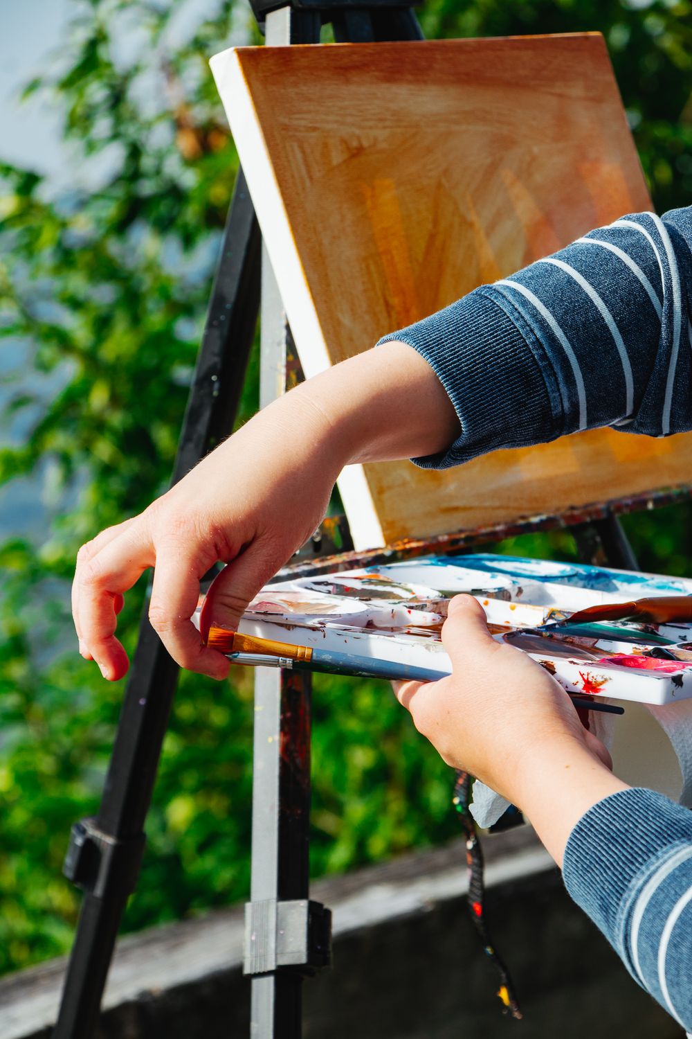 A close-up of hands reaching for a brush from a paint-filled palette, with an easel and sunlit greenery in the background