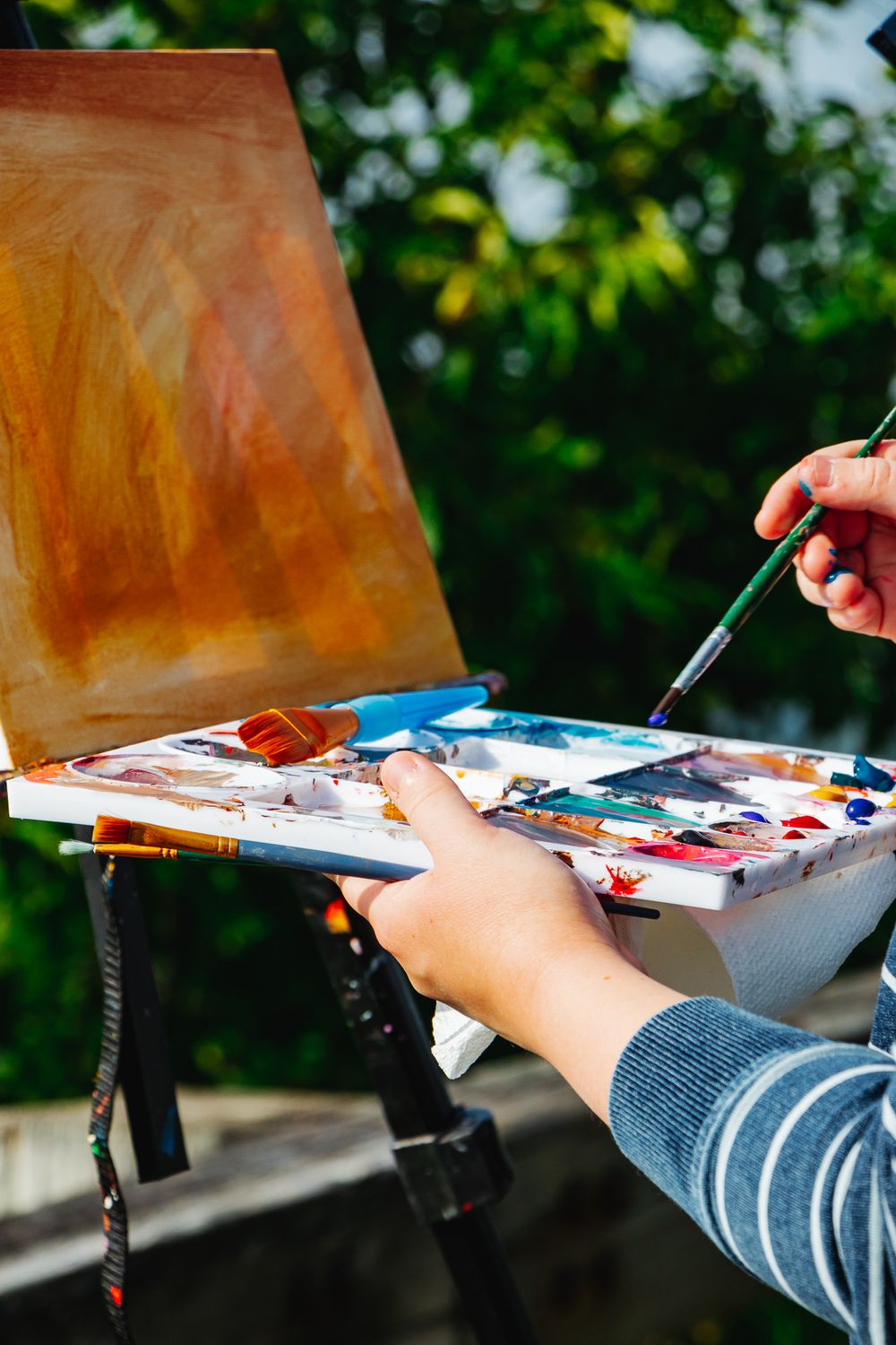 A bright photo of a painter's hands holding a palette full of coloful paints and brushes, in front of a blank canvas set up outdoors.
