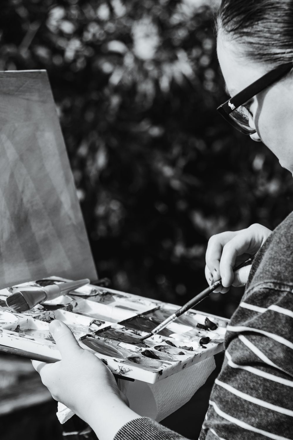 A monochrome image of an artist mixing paint on a palette, with a blank canvas and dappled foliage in the background