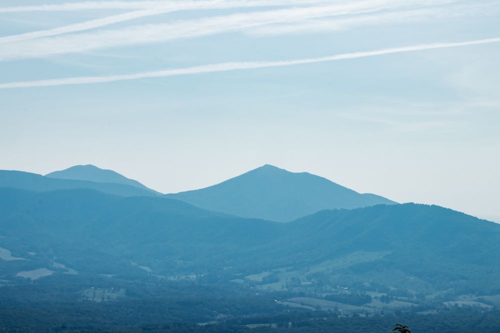 A serene landscape photo of layered Blue Ridge Mountains under a pale blue sky with soft clouds and vapor trails.