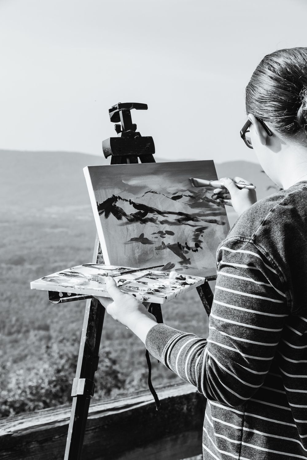 A close-up black and white image of an artist painting a landscape with bold strokes on canvas, holding a palette of mixed paint