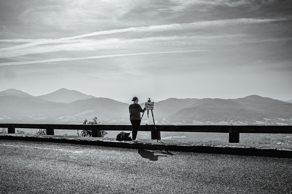 A monochrome photo of an artist painting at a scenic mountain overlook, with layered hills and dramatic skies in the background.