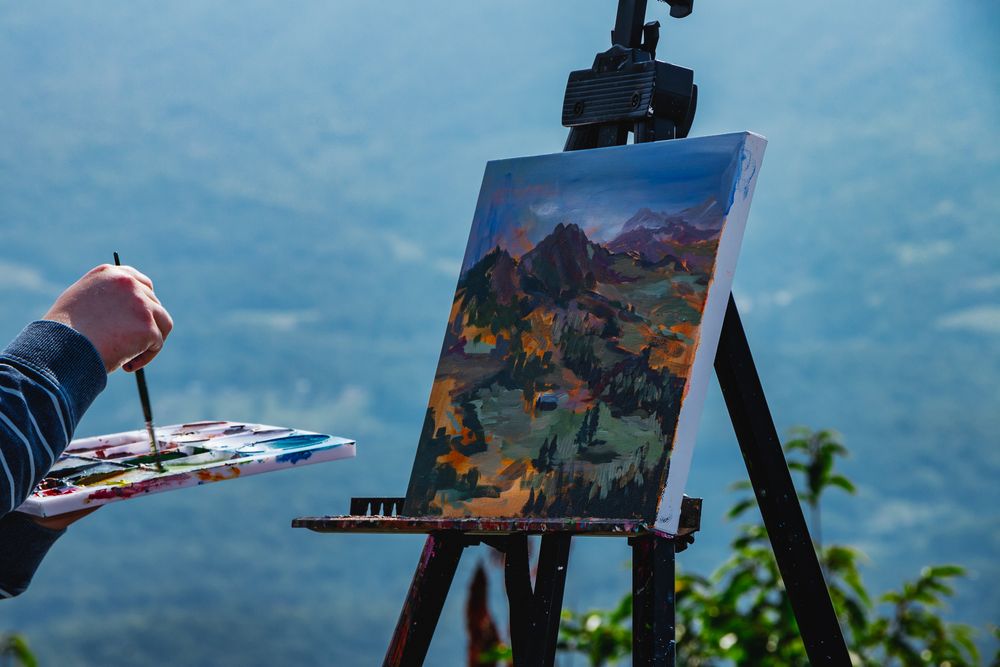 A hand holding a paintbrush and palette mid-stroke, working on a vivid mountain landscape on an easel, with blue-toned hills in the distance