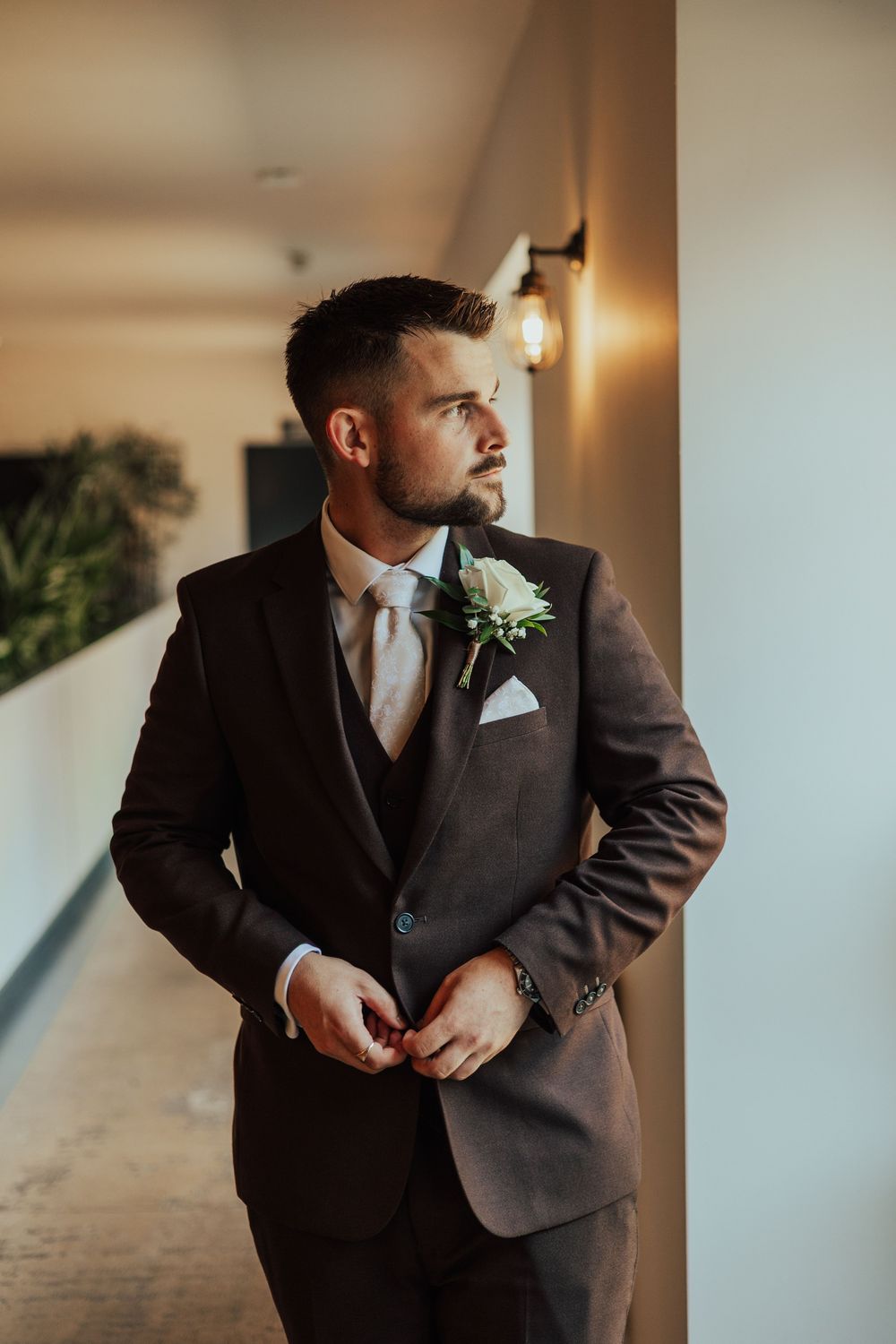 Dapper groom in a dark three-piece suit with boutonniere and light tie, standing in a hallway.