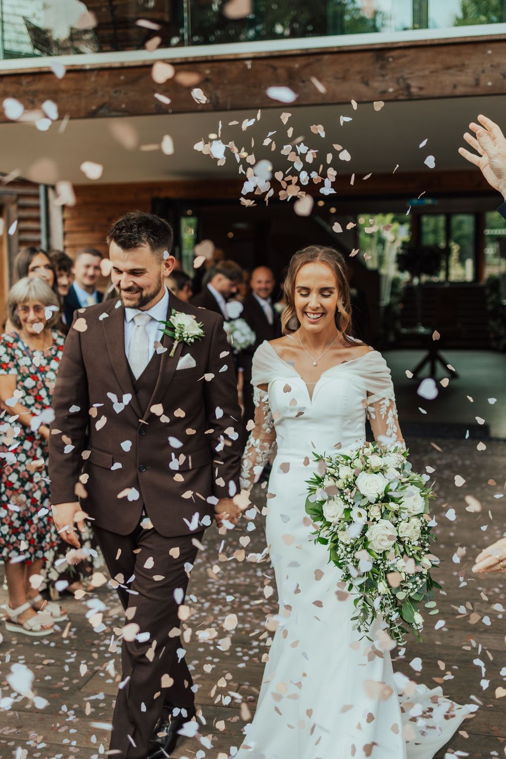 A joyful newlywed couple walks through a shower of confetti petals, bride holding a white floral bouquet, guests celebrating behind them.