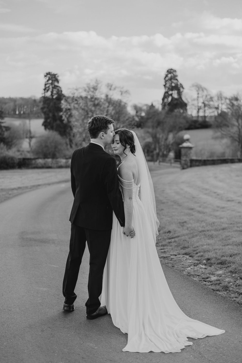 Black and white photo of a bride and groom holding hands on a winding path, with trees and countryside in the background.