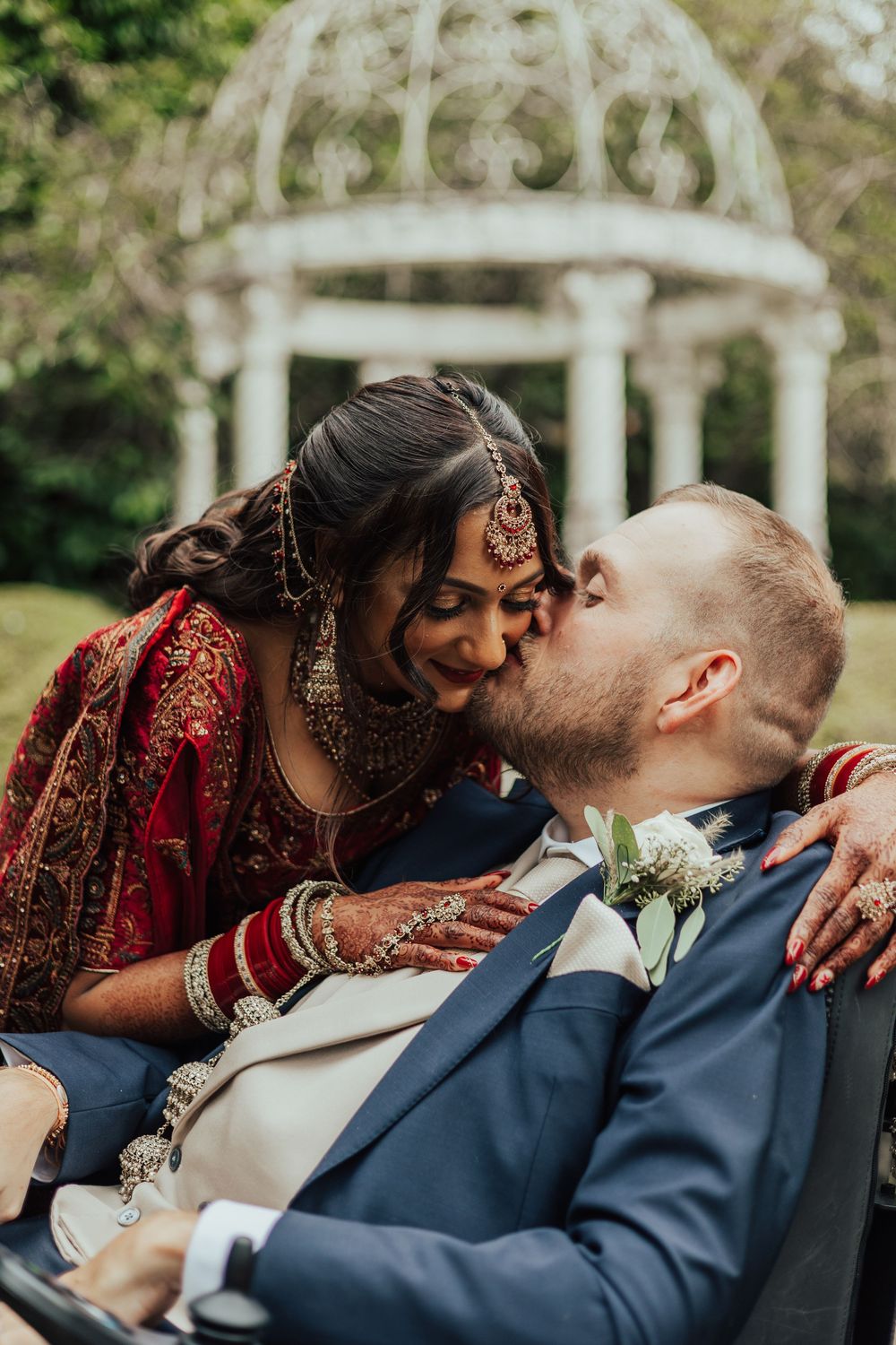 Indian bride in red lehenga kissing groom in blue suit outdoors near white gazebo in lush garden setting.
