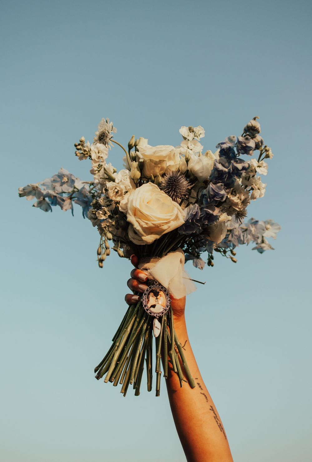 Hand holding up a dried flower bouquet with pale roses and gray foliage against a light blue sky.