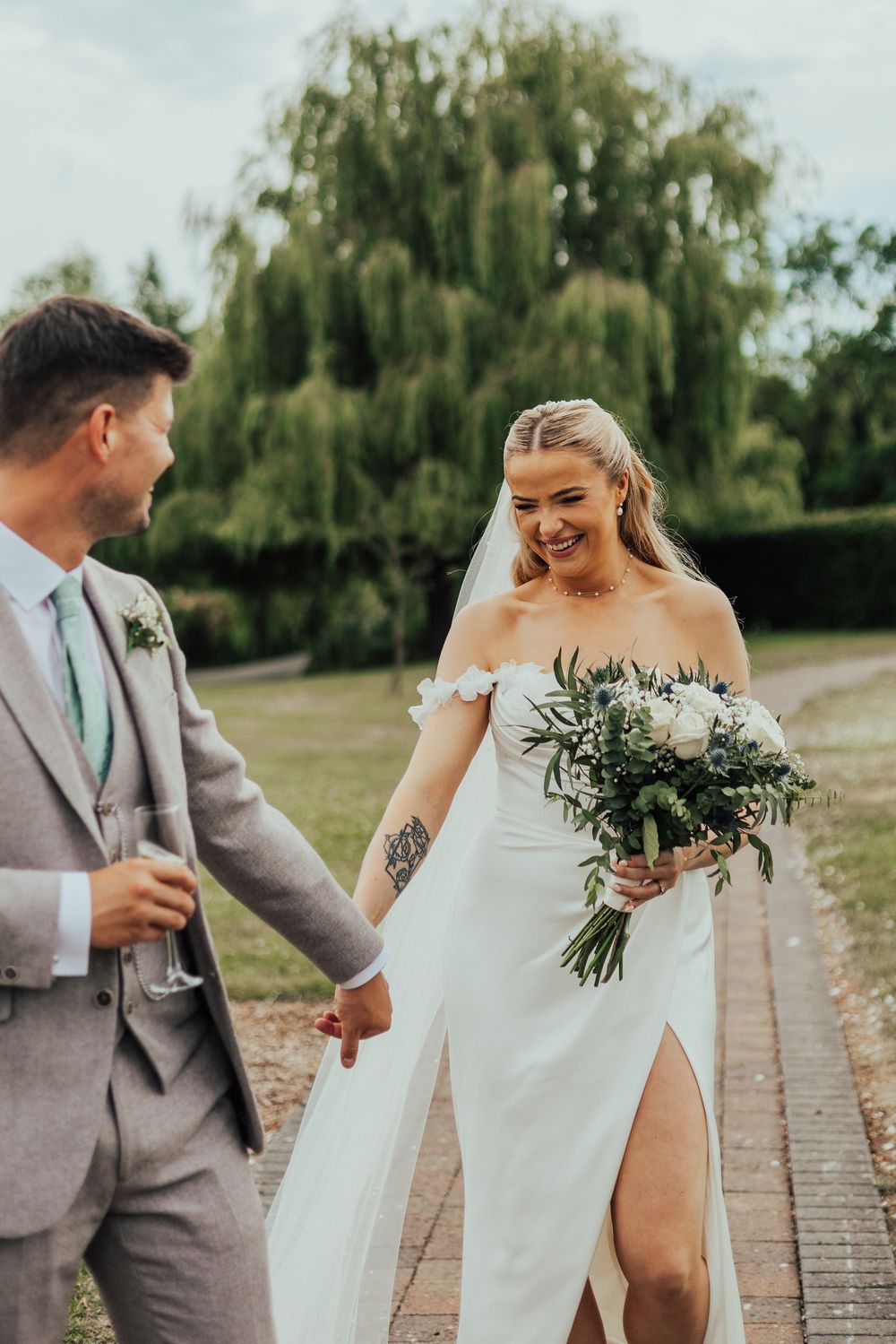 Bride and groom holding hands outdoors, bride in white off-shoulder gown with bouquet, groom in grey suit with champagne.
