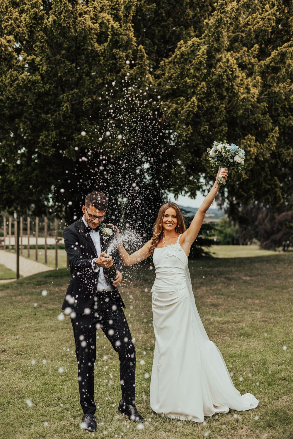 Bride and groom celebrating outdoors, popping champagne bottle with joyful spraying bubbles on green lawn.
