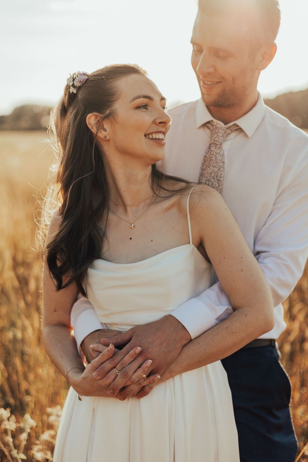 A smiling bride in a white dress embraces her groom in a golden field during a romantic sunset wedding photo.