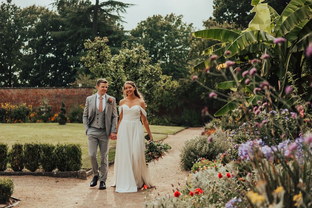 People walking along a garden path surrounded by blooming pink and purple flowers on a sunny summer day.