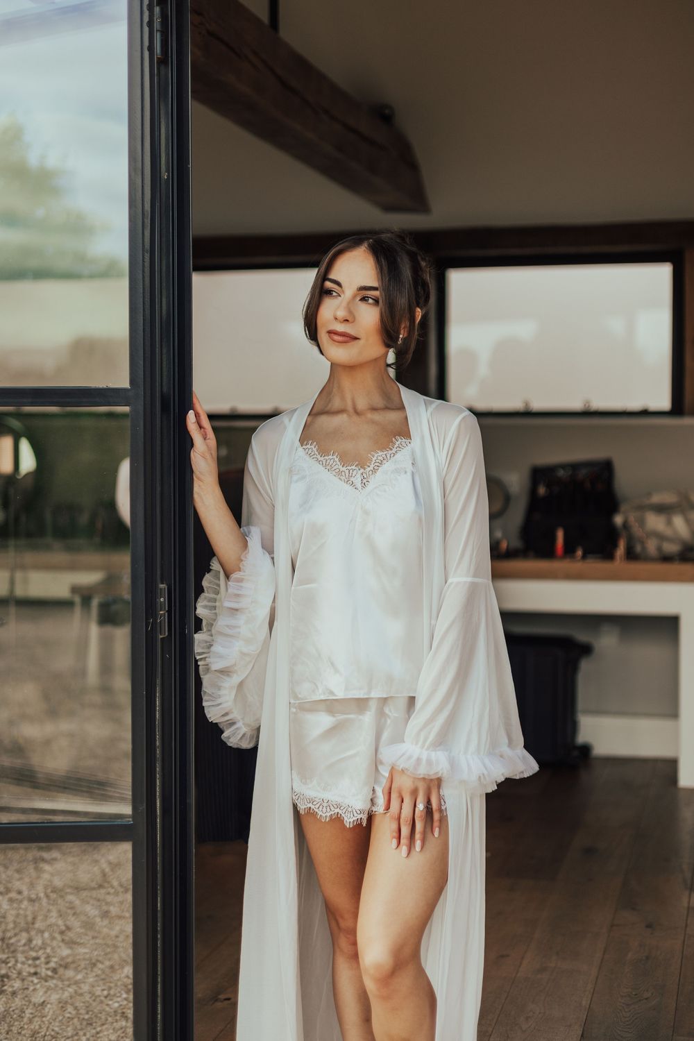 Elegant bride in white satin shorts set and sheer robe poses gracefully in a doorway during bridal morning prep.