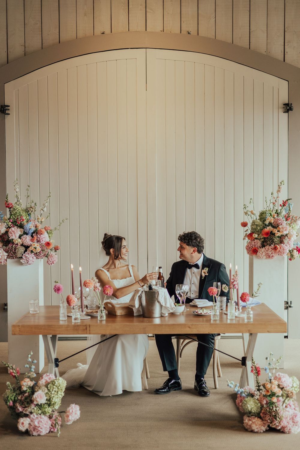 Bride and groom toast at floral-adorned sweetheart table in elegant barn wedding venue with pink blooms.