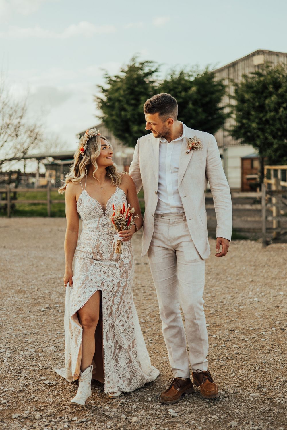 A couple in light-colored wedding attire walks together at a rustic outdoor venue during golden hour.