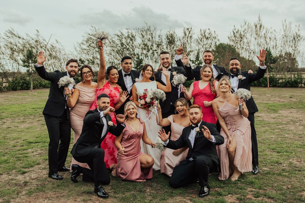 Wedding party members in formal attire pose together on grassy hillside for candid group photos during outdoor celebration.