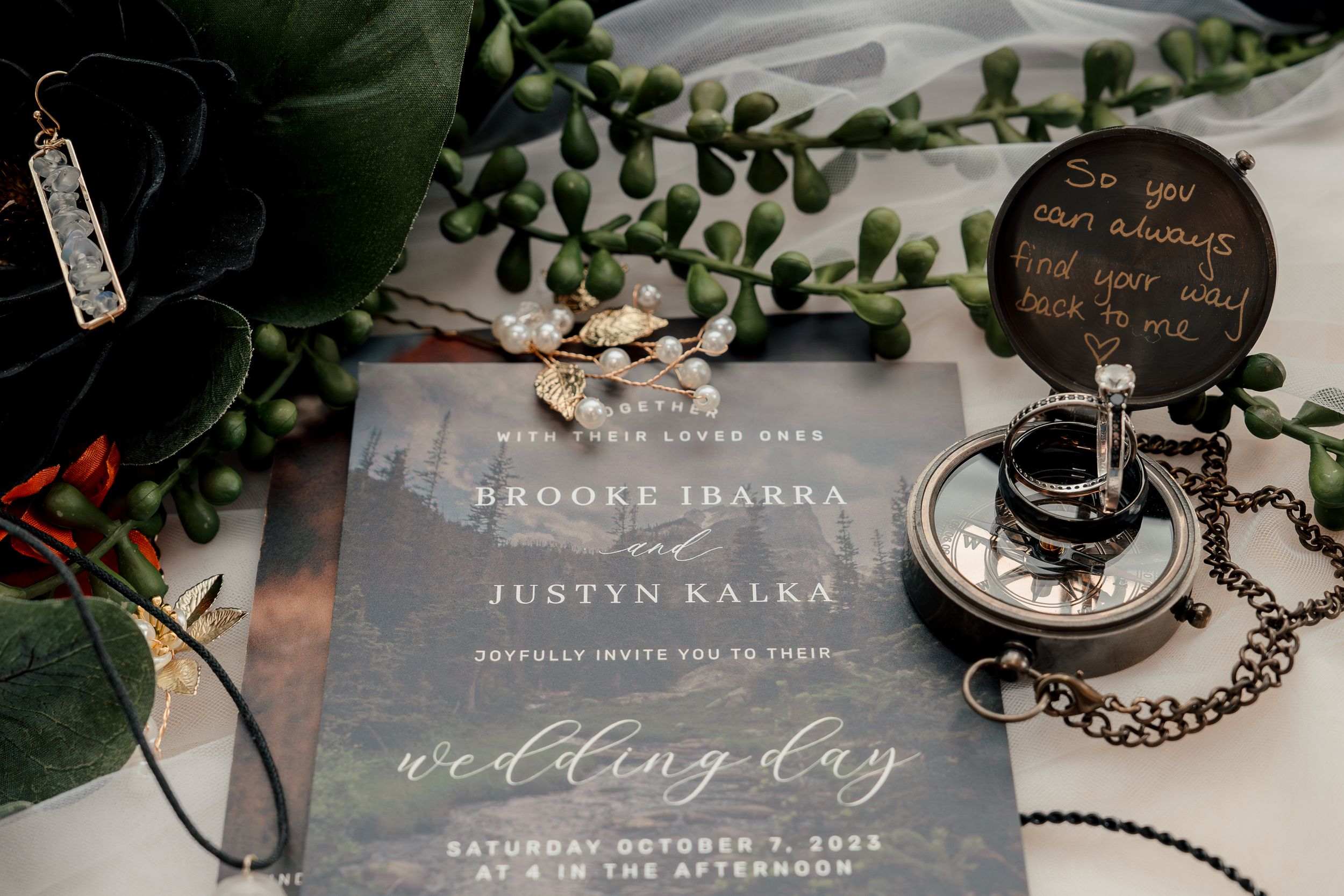 A vintage pocket watch lies beside a wedding invitation with delicate greenery and floral decorations.
