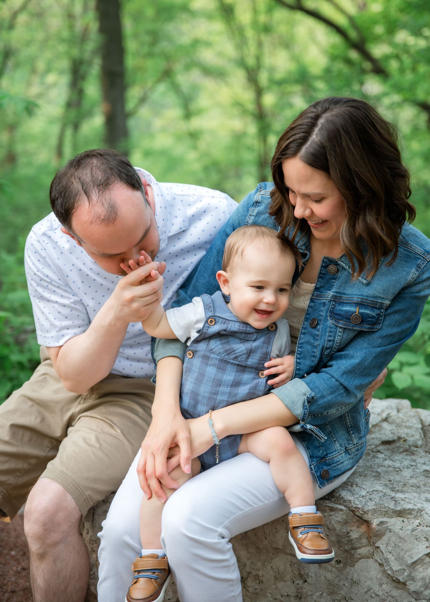 Family sits together on a rock in a lush green forest setting, sharing a tender candid moment together on a summer day.