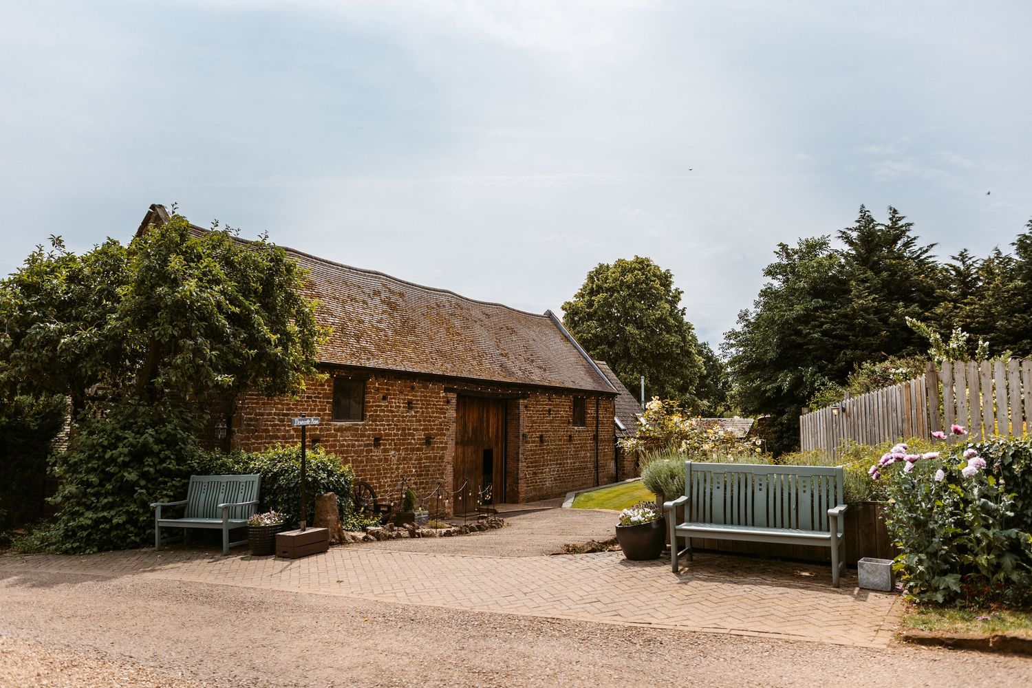 A row of rustic brick farm buildings and barns with thatched roofs set against a rural countryside landscape.