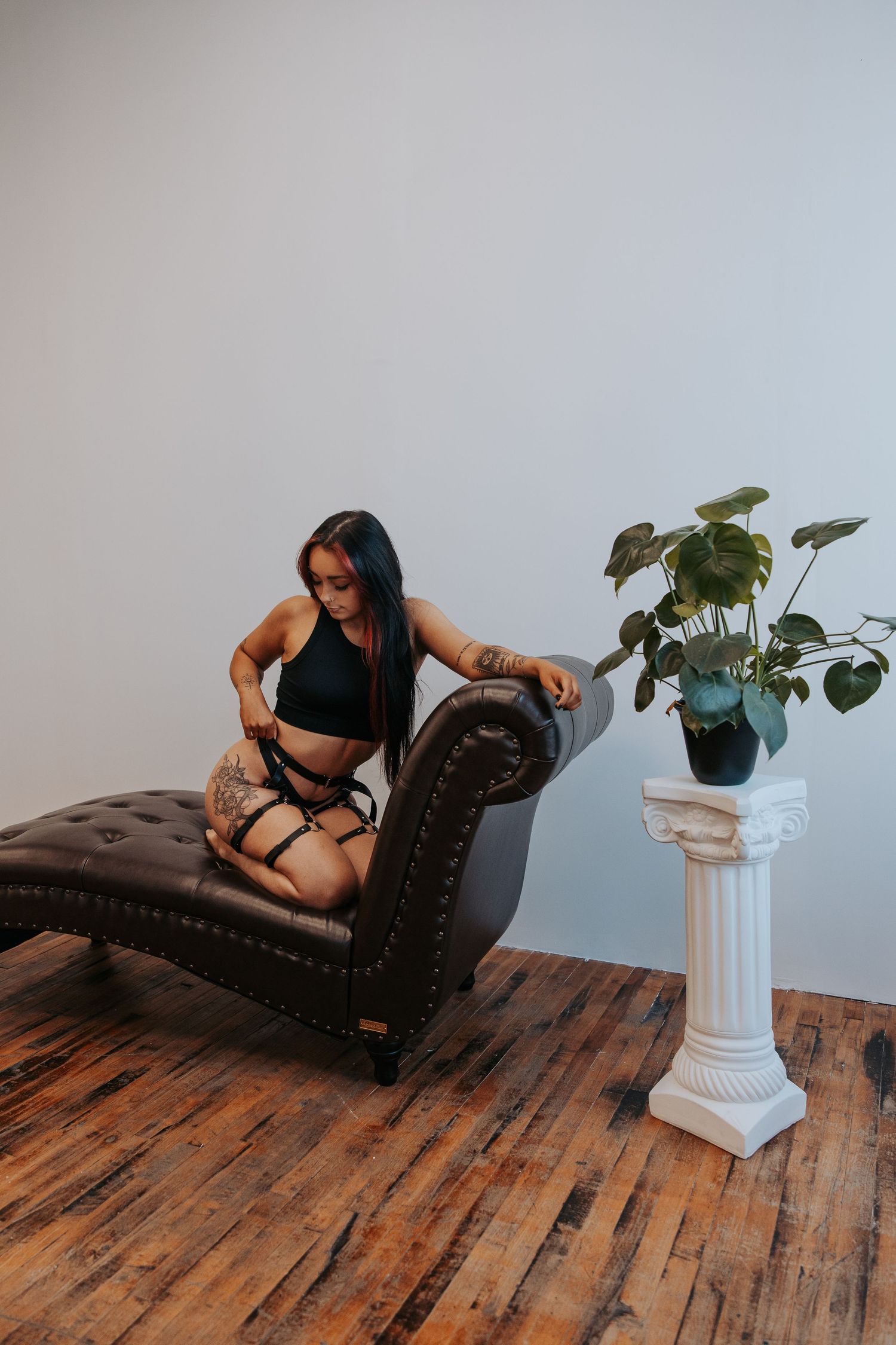 A person in black attire poses on a tufted leather chaise lounge next to a potted plant.