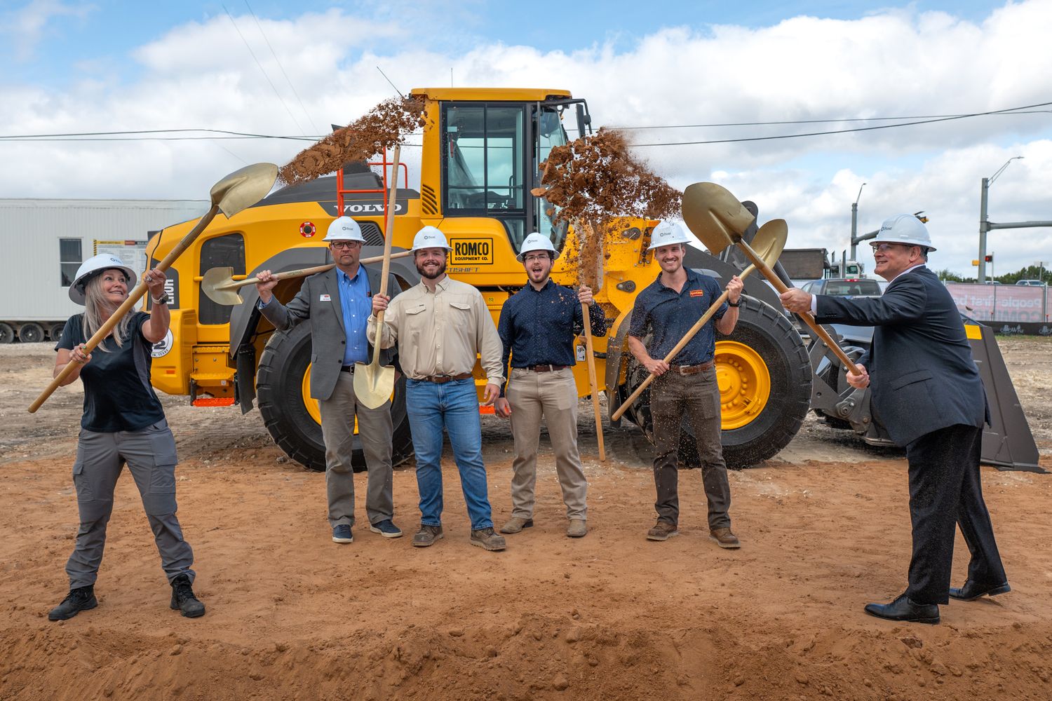 Six people in hard hats pose with shovels at a groundbreaking ceremony in front of a yellow front loader.