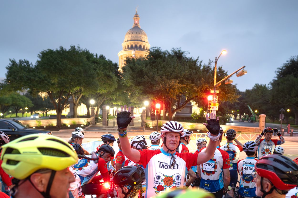 Cyclists gather near Texas State Capitol at dusk for a charity bike ride, with a rider in red raising arms in celebration.