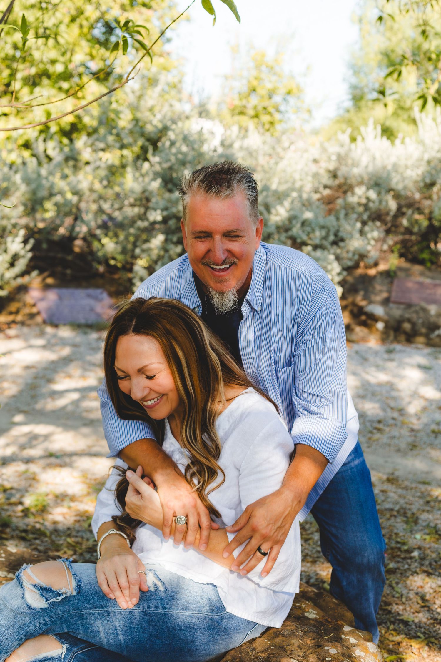 A couple shares a joyful embrace while sitting outdoors in a sunlit natural setting with trees in the background.