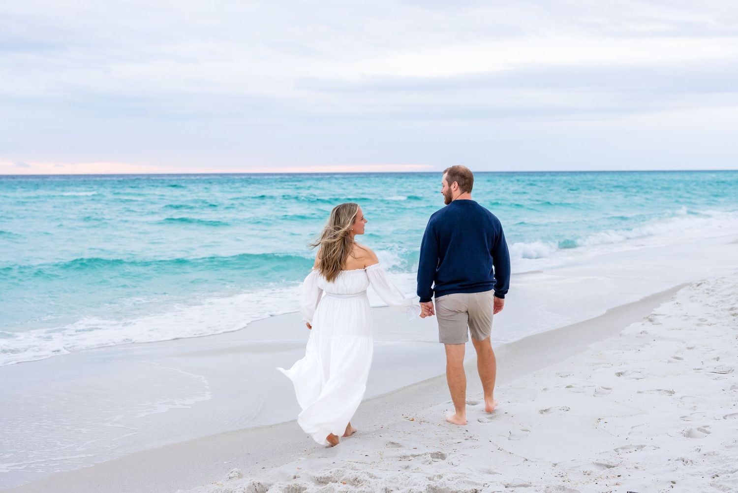 A couple walks hand in hand along a pristine white sand beach with turquoise ocean waters in the background.