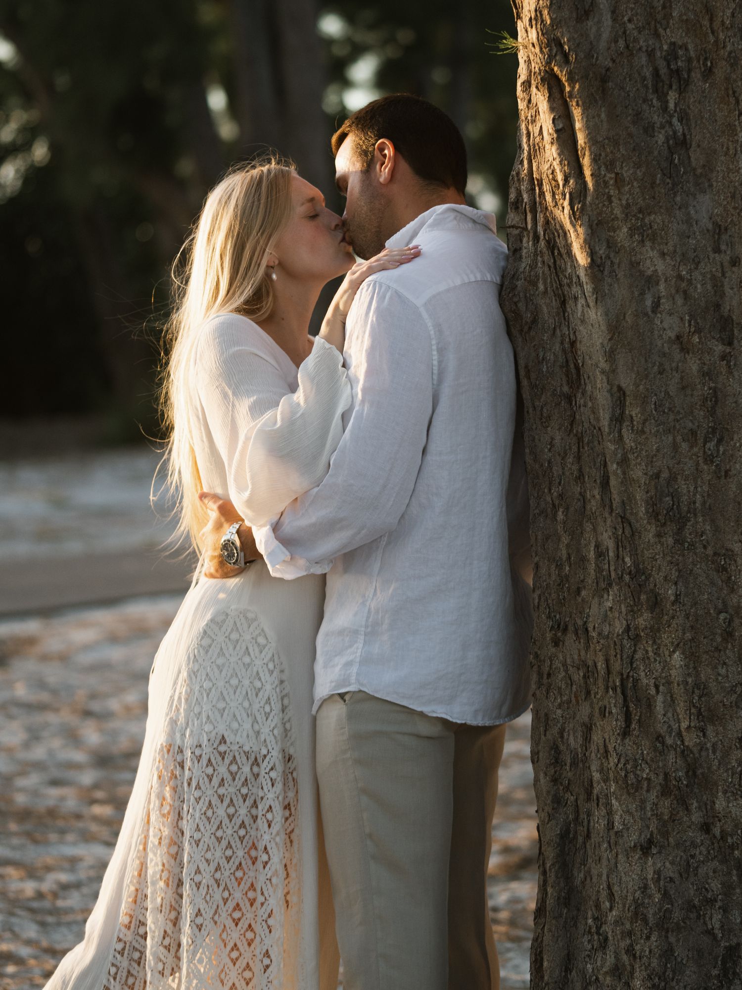Intimate moment captured between couple in white outfits against a tree.