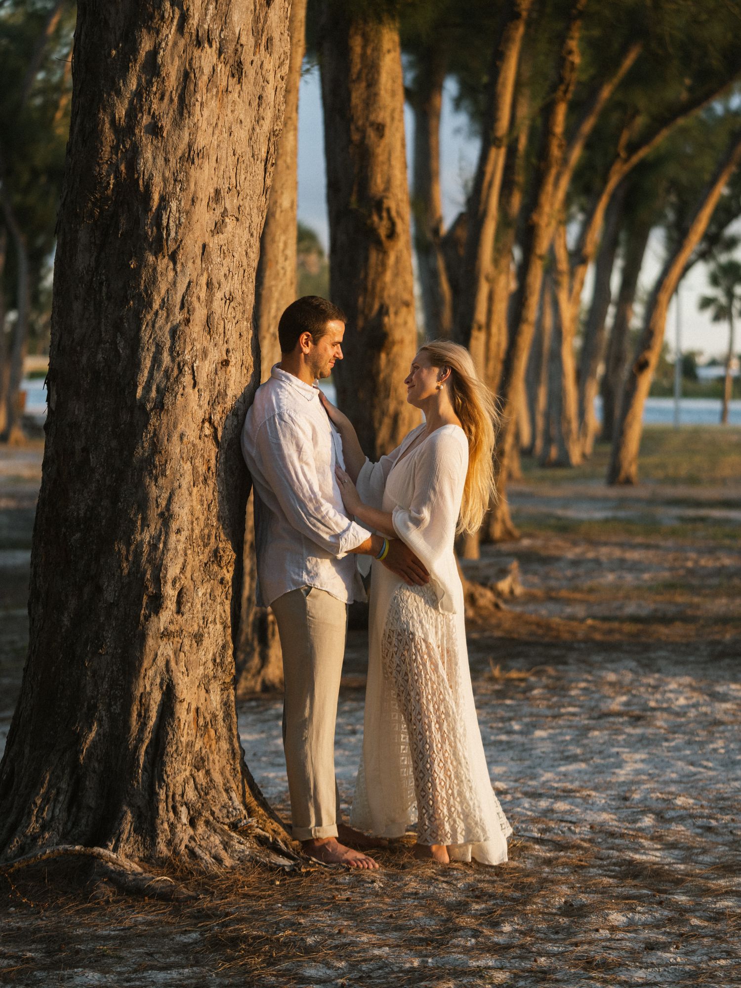 Romantic couple in white outfits standing between tall trees on a sandy beach at golden hour.
