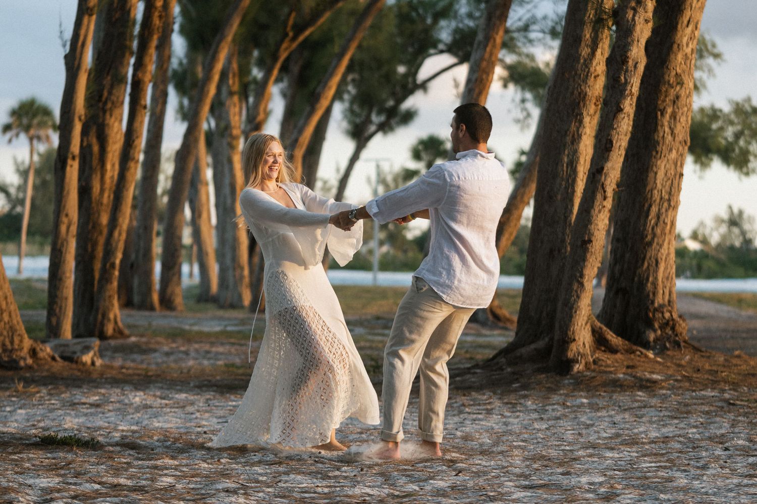 Couple in white outfits walking together through a scenic tree-lined path at sunset.