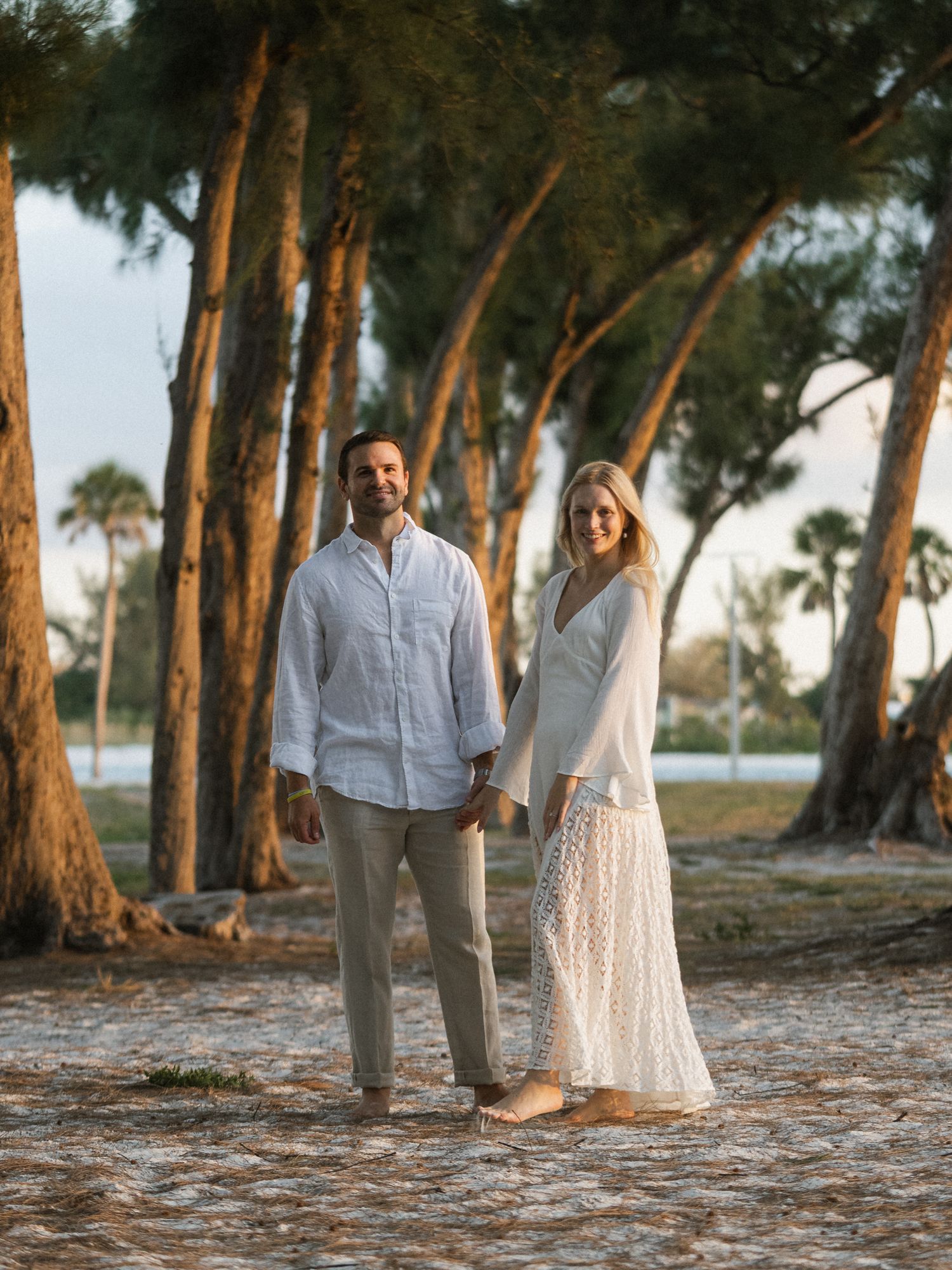 Couple in coordinated white outfits standing among tall pine trees at sunset.
