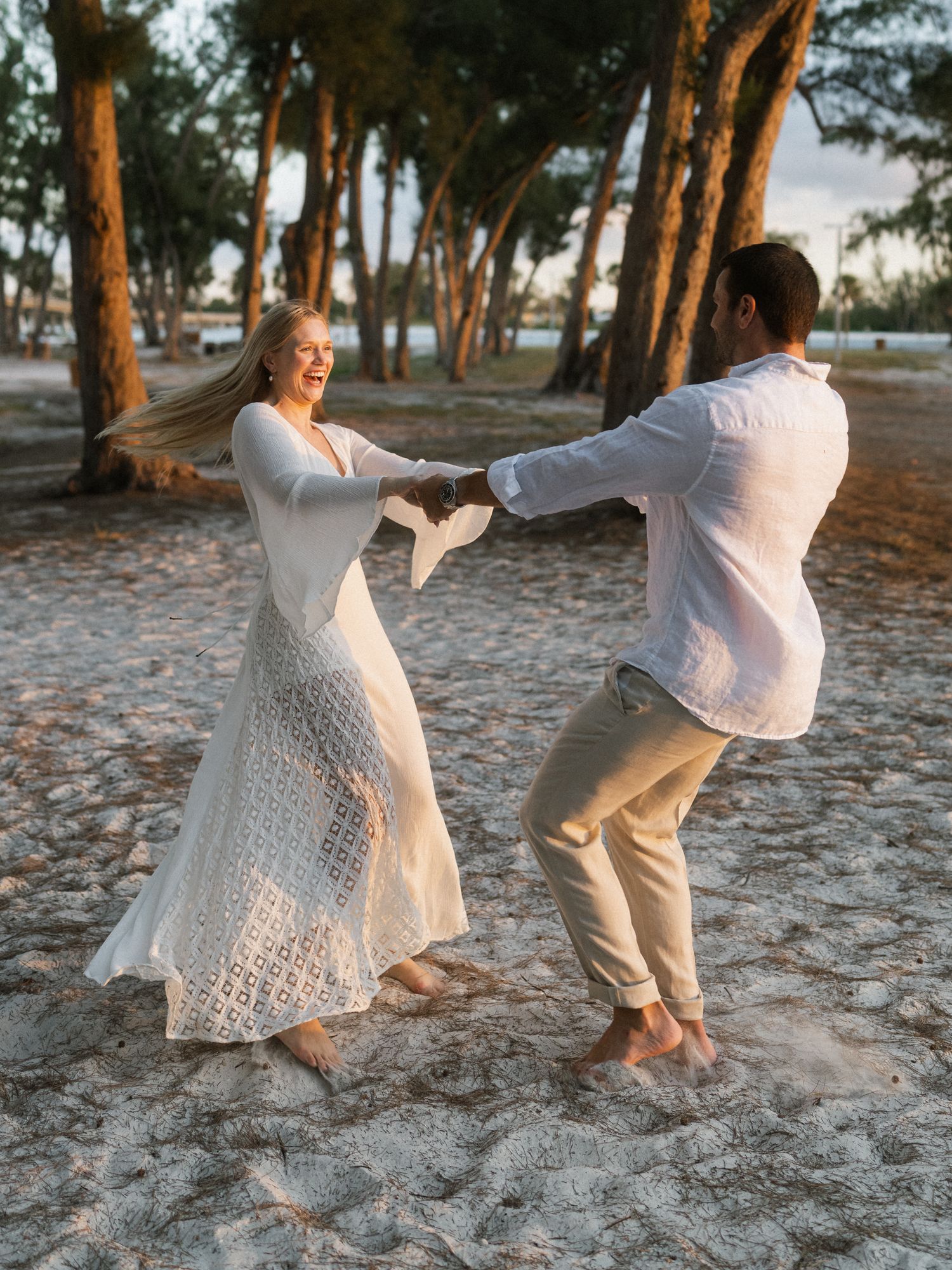Two people in white outfits dance playfully on a sandy beach surrounded by pine trees at sunset.