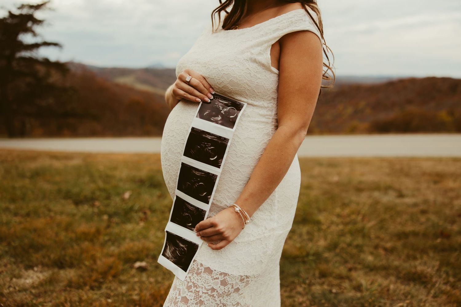 A pregnant woman in a white lace dress holds ultrasound photos against her baby bump in an outdoor field in The Blue Ridge Mountains.
